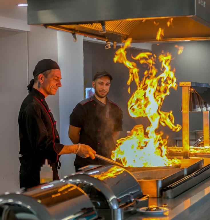 Šefpavārs, gatavojot ēdienu ar liesmām, kopā ar palīgu, kas vēro procesu. Aiz viņiem redzama moderna virtuve ar gatavošanas iekārtām.