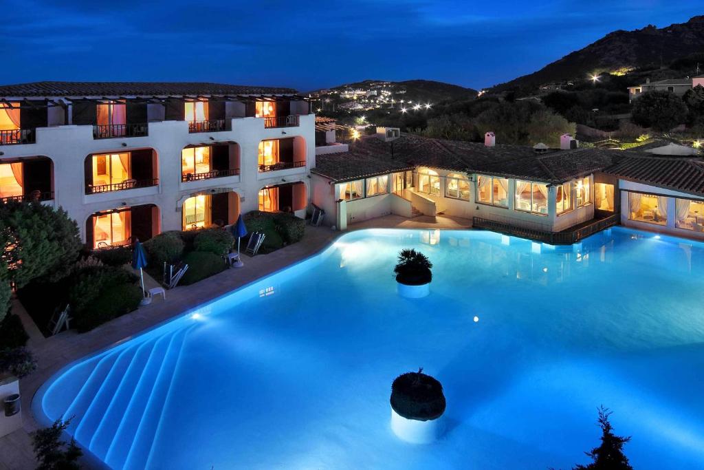 Nakts skats uz viesnīcu ar zilu baseinu un apgaismotām terasēm, kas rada mierīgu atmosfēru. Apkārt esošie pakalni un debesu apgaismojums piešķir ainavai burvību.