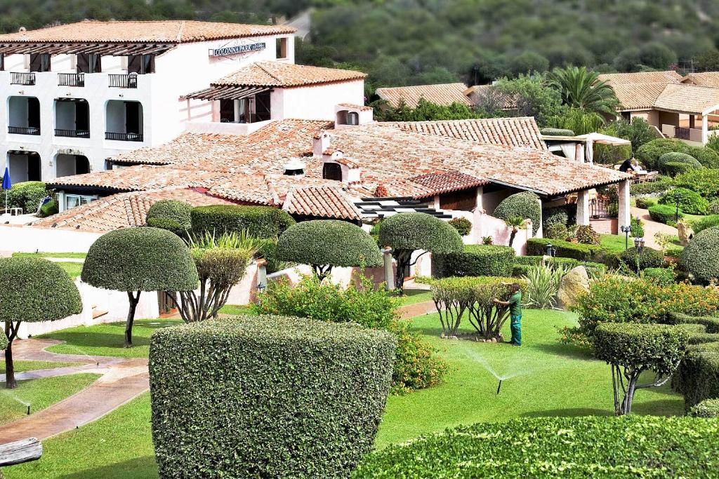 Skaists dārza skats ar labi koptiem krūmiem un zālienu, kurā redzams darbinieks, kas laista augus. Fons veido arhitektūras elements ar tradicionālu jumtu un balkoniem.