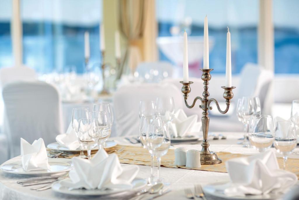 Eleganti iestatīts galds ar baltiem galdautiem, stikla traukiem un sudraba svečturiem. Gaisīga atmosfēra pie logiem ar skatu uz ūdeni.