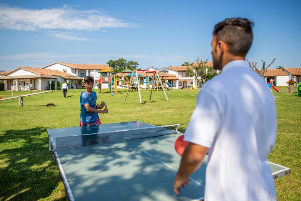 Divi jaunieši spēlē galda tenisu atpūtas centrā, kamēr apkārt ir zaļa zāle un bērnu rotaļu laukums. Fons ir mājas ar sarkanām jumta flīzēm un skaidrs debesis.