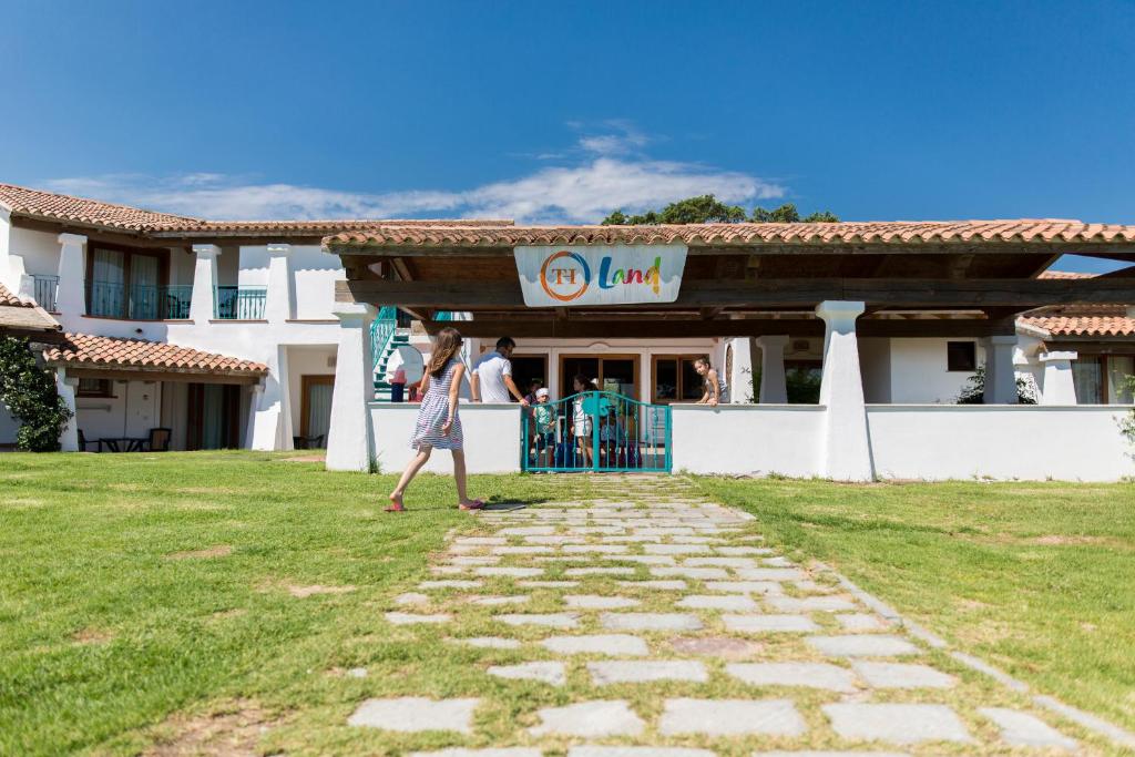 Mājas ēka ar baltiem sienām un jumtu no sarkanām flīzēm, kas atrodas zaļā pļavā. Priekšplānā redzama meitene, kas dodas uz ieeju, kur pulcējas cilvēki. Skats uz zilo debesis.