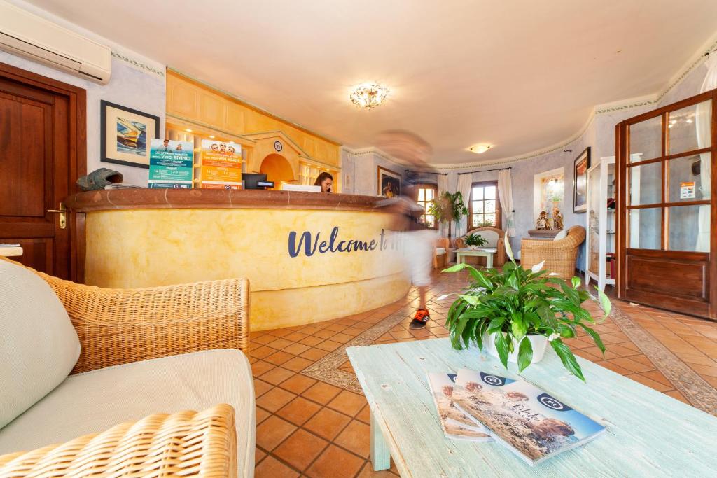 Viesnīcas reģistratūra ar siltu atmosfēru un augu dekorācijām, kur apmeklētājs uzrunā viesmīli pie letes. Interjers ir gaišs un viesmīlīgs.