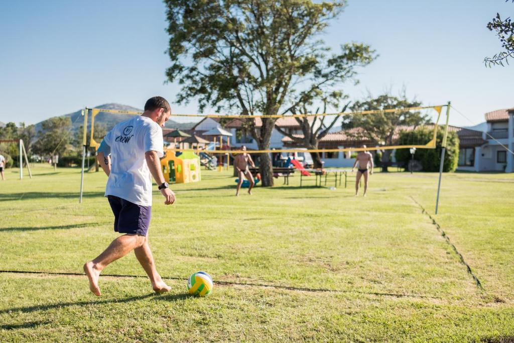 Volejbola spēle saulainā dienā, kurā redzams vīrietis, kas iet uz laukuma ar bumbu, kamēr citi spēlētāji ir fona.