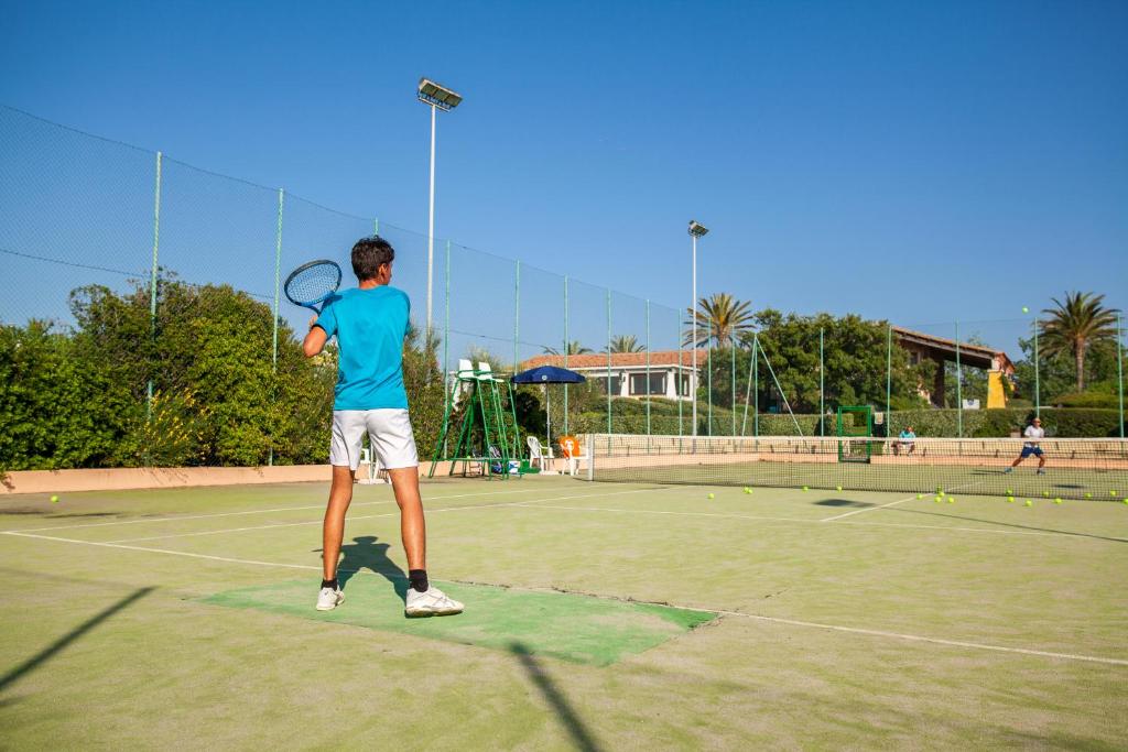 Jaunietis spēlē tenisu uz laukuma, saulainā dienā, apkārt ir zaļumi un citi spēlētāji.