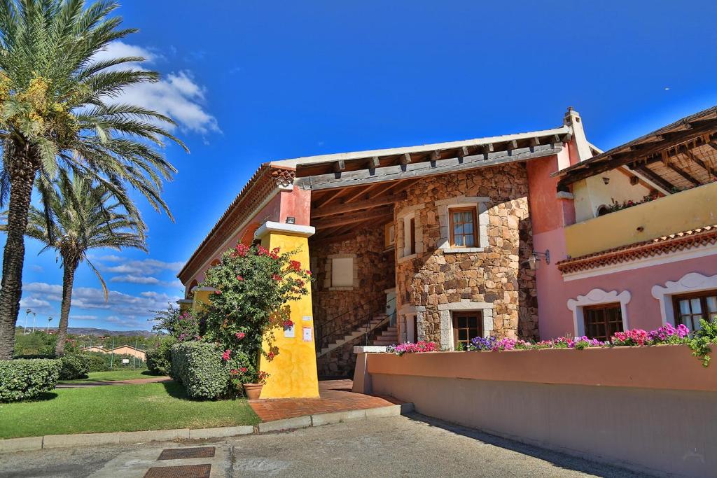 Krāsaina māja ar akmens fasādi un ziediem priekšā, ko ieskauj palmas un skaidrs debesis. Ideāla ainava relaksācijai un brīvā laika pavadīšanai.