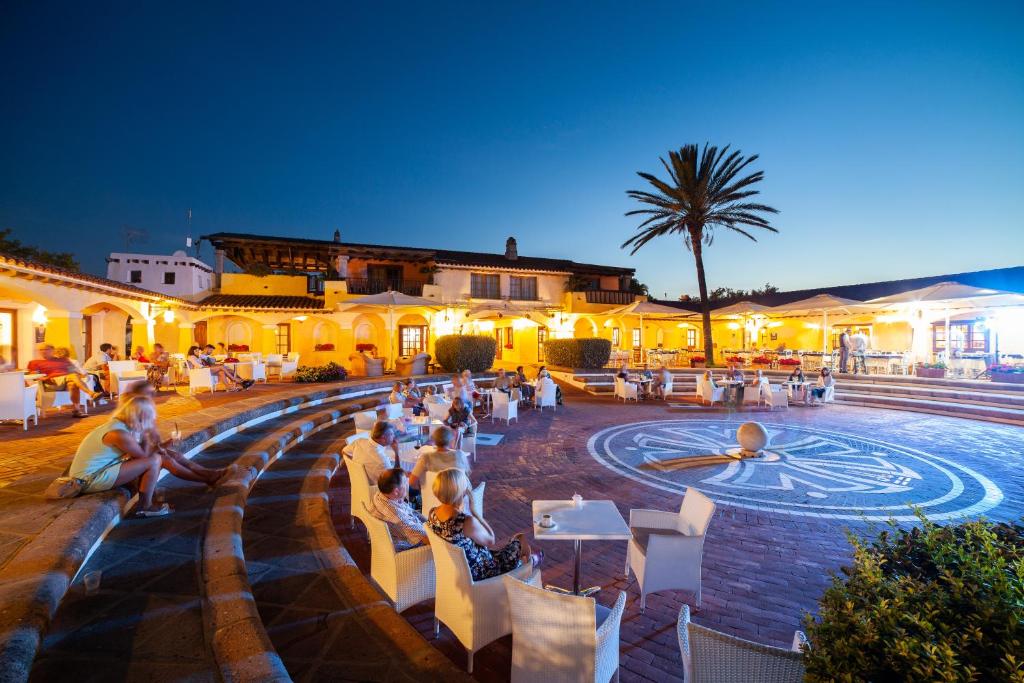 Vakara ainava ar cilvēkiem, kas atpūšas pie ērtām sēdvietām terasē, apgaismotā vidē ar palmām un tradicionālo arhitektūru.