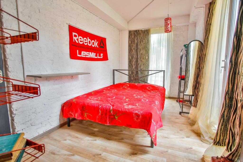 Mūsdienīga guļamistaba ar sarkanu gultas veļu, Reebok plakātu un vieglām aizkariem. Telpā ir koka grīda un spogulis, radot gaišu un elegantu atmosfēru.