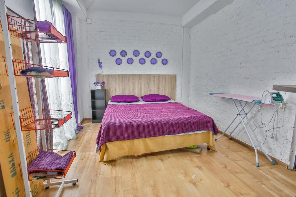 Mūsdienīgs guļamistabas interjers ar divguļamo gultu, violetiem akcentiem un koka grīdu. Istabā ir arī drēbju plaukts un gludināšanas dēlis.