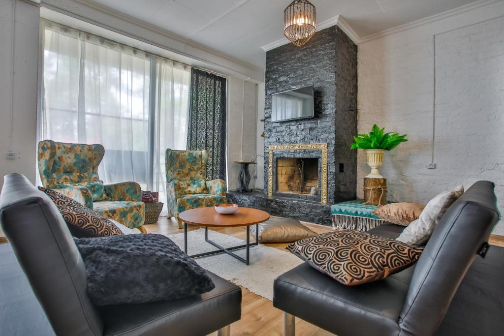 Mūsdienīgs viesistabas interjers ar melnu akmens kamīnu, gaišām logu žalūzijām un komfortablām krēslām. Iekārtots ar dekoratīviem spilveniem un elegantu galdu, radot siltu un viesmīlīgu atmosfēru.