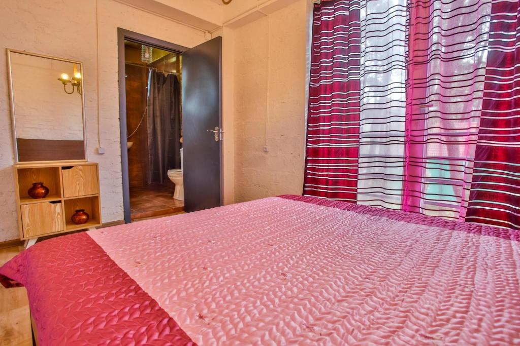 Guļamistaba ar rozā gultas veļu un košām aizkaru strīpām, redzama arī spoguļa un plauktu kombinācija. Durvis ved uz vannas istabu.