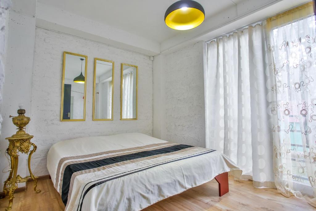 Mūsdienīga guļamistaba ar gultu, trīs spoguļiem un gaišiem logiem, kas nodrošina dabisko apgaismojumu. Interjers ir vienkāršs, bet elegants, ar stilīgu lampu un dekoratīvu statīvu.