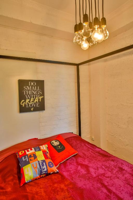 Skaista guļamistaba ar sarkanu gultu, spilventiņiem un stilīgu lampu kaskādi. Uz sienas ir iedvesmojoša plakāts ar uzrakstu "DO SMALL THINGS WITH GREAT LOVE".