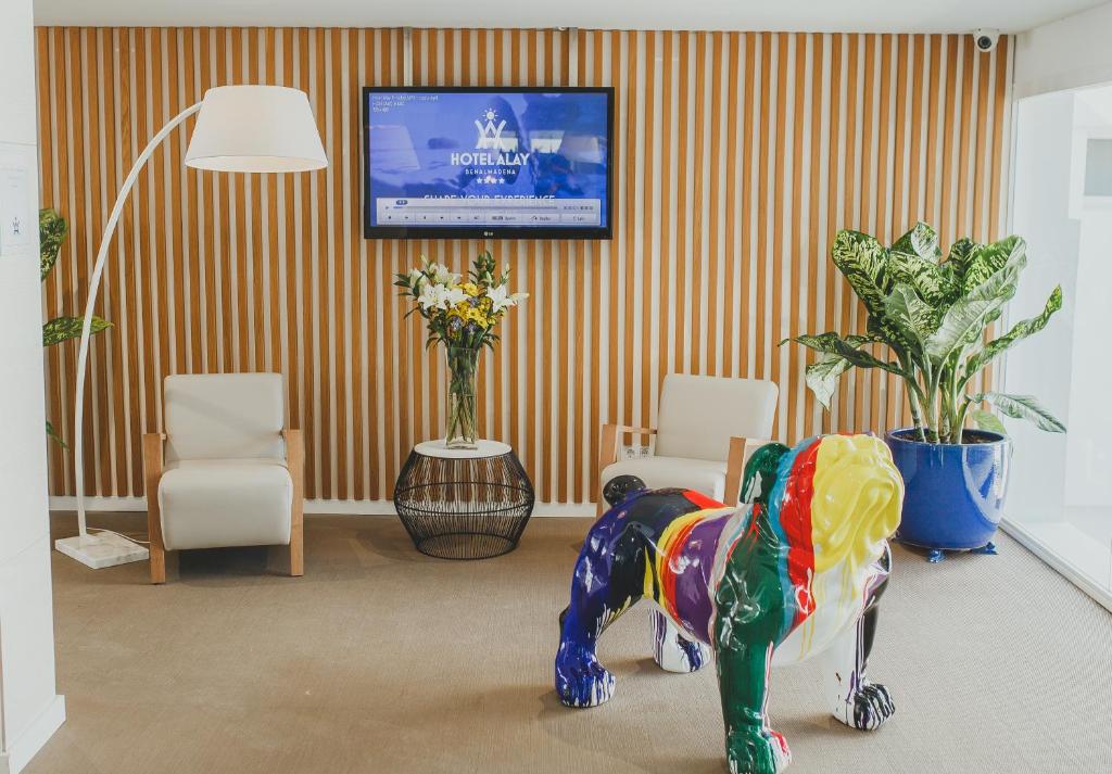 Krāsains lauva skulptūras attēls viesnīcas uzgaidāmajā telpā, kurā ir moderns dekors ar koka sienām, elegantu apgaismojumu un ziediem. Fons ietver televizoru ar viesnīcas nosaukumu un logo.