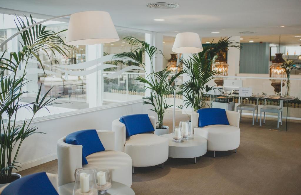 Mūsdienīga atpūtas zona ar baltām krēslām un ziliem spilveniem, ko ieskauj zaļi augi un gaiša apgaismojuma elementi. Eleganta un komfortabla atmosfēra.