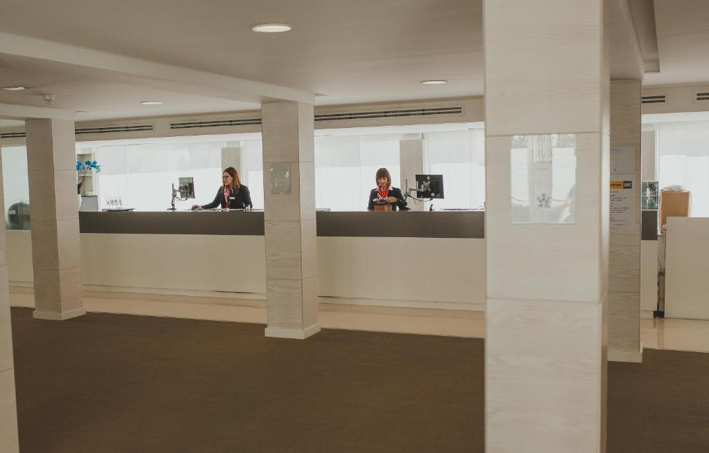 Recepcija modernā viesnīcā ar divām darbiniecēm, kas strādā pie galda, un gaišu, plašu telpu. 