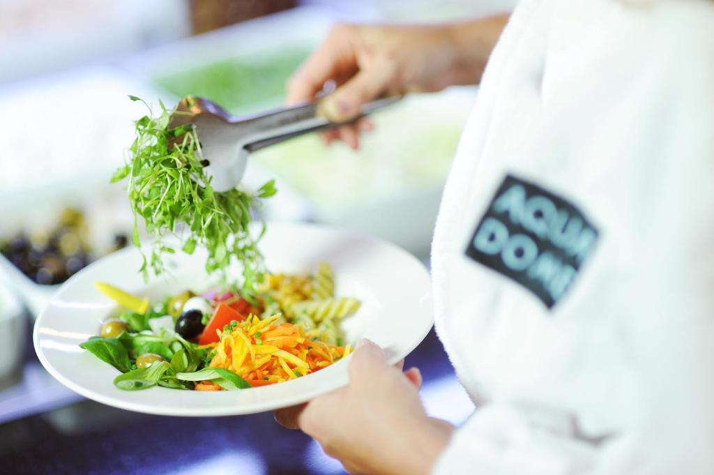 Svaigā salātu pasniegšana ar dažādiem dārzeņiem, zaļumiem un olīvām, kamēr persona tērpta halātā.