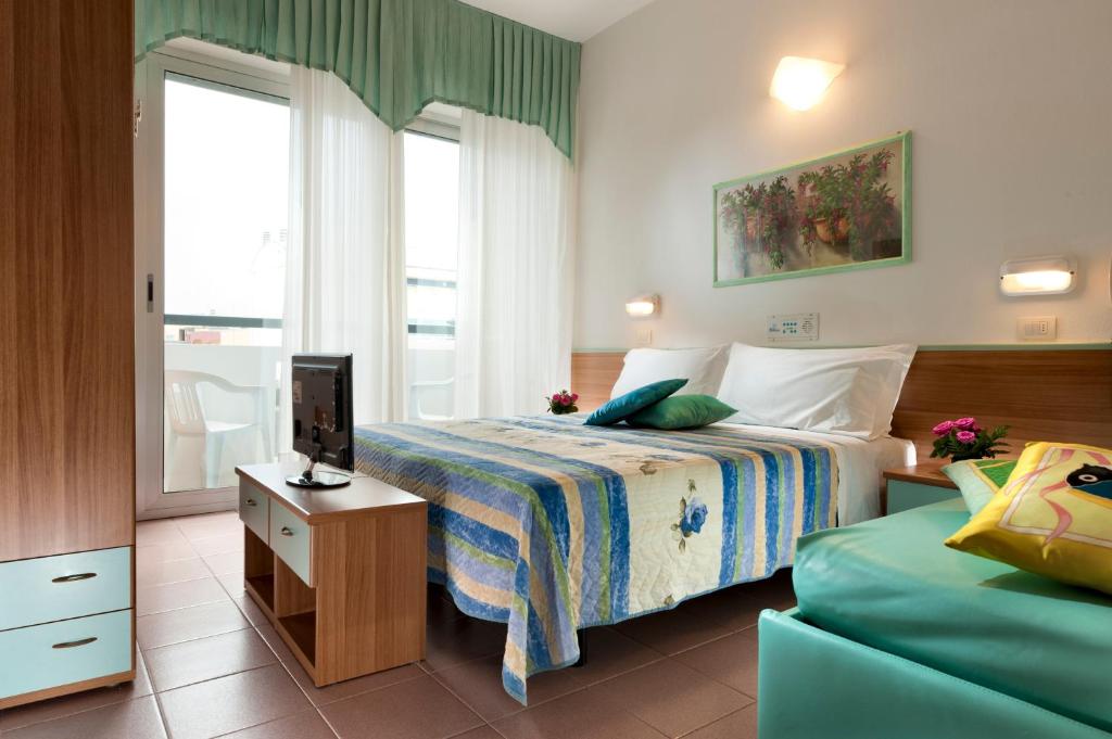 Mūsdienīga viesnīcas istaba ar divguļamo gultu, televizoru un balkonu. Interjers ir gaišs ar krāsainu gultas veļu un dekoratīviem spilveniem, radot mājīgu atmosfēru.