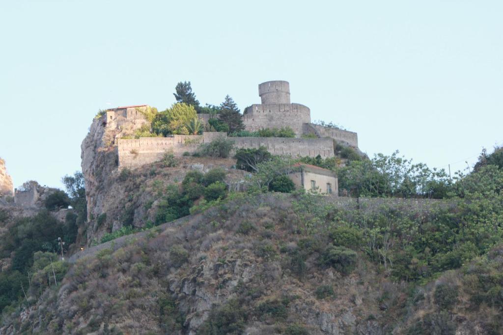 Kalna cietoksnis ar apaļu torni un mūriem, ieskauts zaļiem kokiem un krūmiem. Fons ir skaidrs debesis.