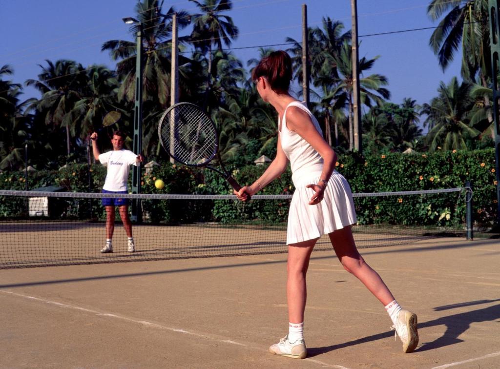 Tennisa spēles mirklis ar diviem spēlētājiem, no kuriem viens gatavojas uzsist bumbu, bet otrs ir ieņēmis gatavības pozīciju. Fons ir tropiska vide ar palmām.