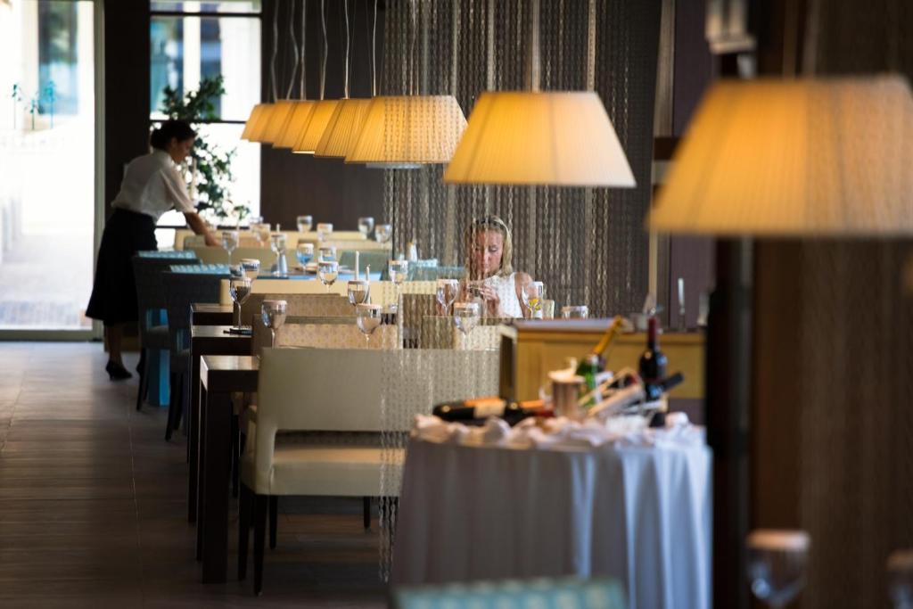 Elegants restorāns ar izsmalcinātu apgaismojumu un galdiem, kas sakārtoti gaidīšanai uz viesiem. Pamatā ir gaismas un moderns interjers, kā arī viesmīlis, kas apkalpo.