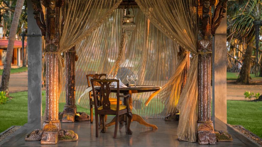 Romantiska ēdamzona ar elegantām koka mēbelēm, apgaismota ar siltu gaismu un rotāta ar caurspīdīgām aizkarām, kas rada intīmu atmosfēru. Apkārt zaļojoša daba un palmām, veidojot mierīgu un pievilcīgu vidi.