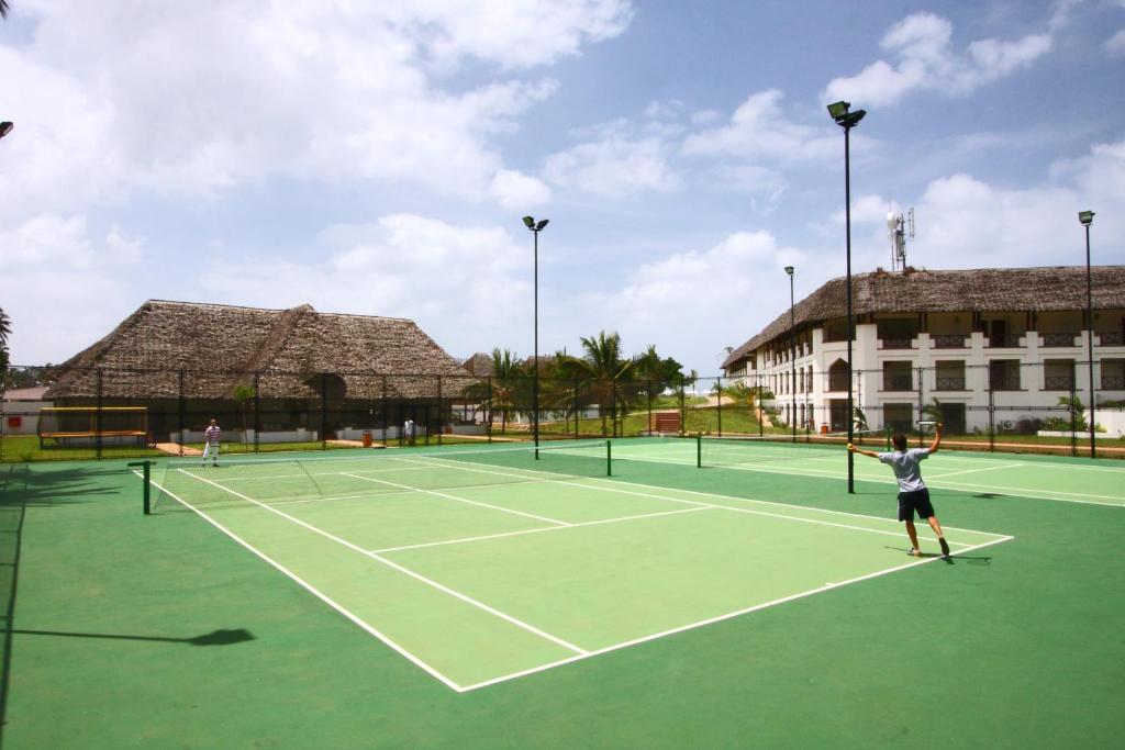 Tennisa laukums ar diviem spēlētājiem, kas spēlē, un tradicionālu ēku fonā. Zilās debesis un saulains laiks rada patīkamu atmosfēru.