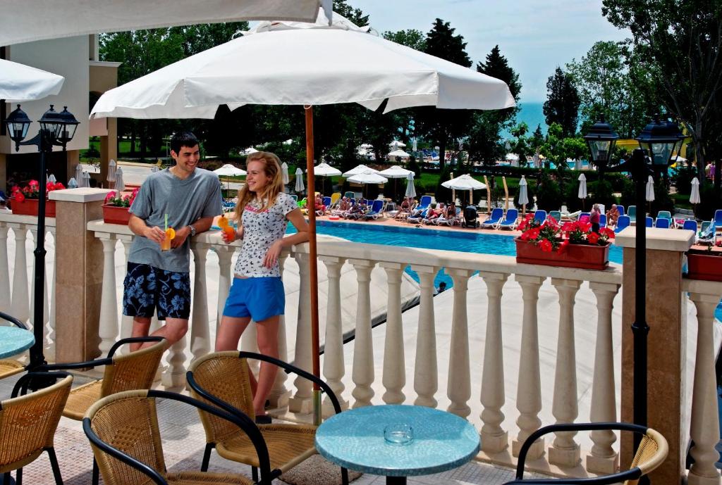 Jaunais pāris izbauda kokteiļus pie baseina, kamēr saule spīd un apkārt ir brīvdienu atmosfēra. Fons ietver peldēšanas baseinu un sauļošanās krēslus.