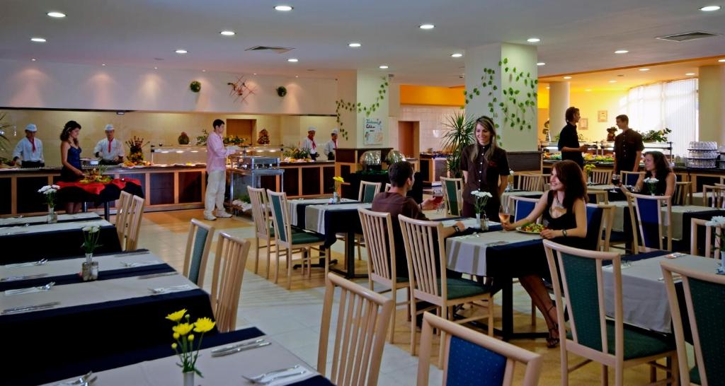Restorāna iekšskats, kur viesi bauda maltītes un apkalpojošais personāls piedāvā ēdienus. Telpa ir gaiša un moderni iekārtota, ar ziediem uz galdiem un bufetes staciju.