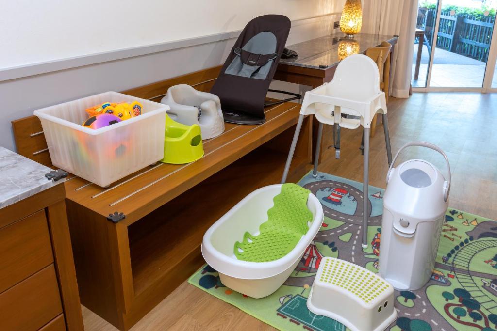 Bērnu aprīkojuma un rotaļlietu stūris ar krēslu, vannu un plastmasas kasti, kas piepildīts ar krāsainām rotaļlietām. Vieta ir gaiša un mājīga ar paklāju, kas attēlo ceļu un automašīnas.