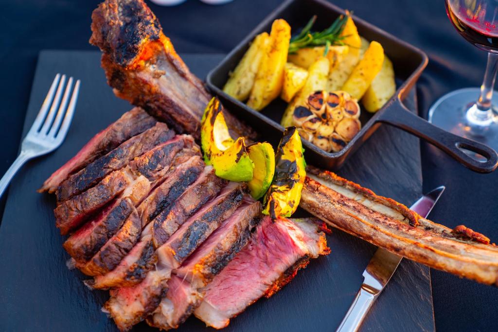 Grilēta gaļa ar dārzeņiem un garšvielām, pasniegta uz melna akmens šķīvja kopā ar ceptiem kartupeļiem un sarkanvīnu.