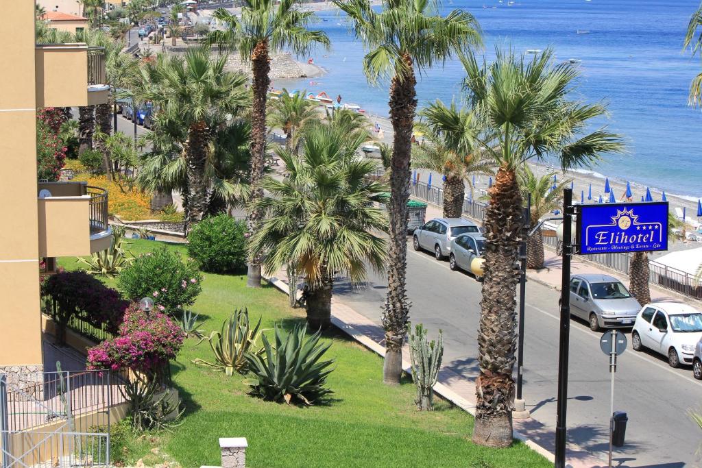 Pludmales ainava ar palmām, ziediem un viesnīcas signālu, kas atrodas netālu no jūras. Viegla vasaras atmosfēra un ceļojuma noskaņa.