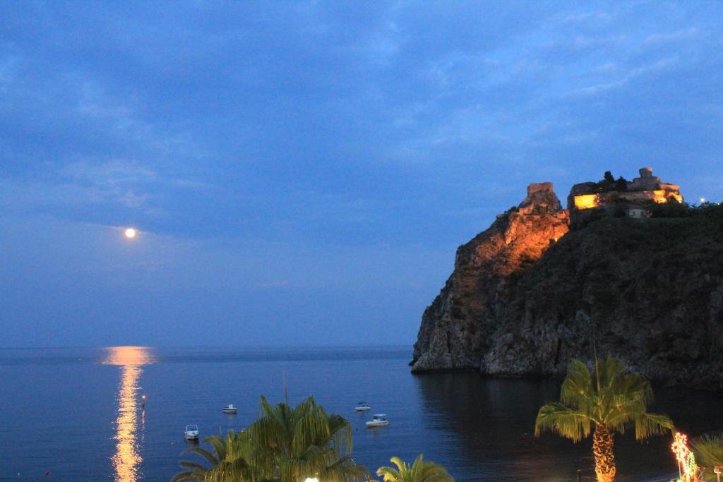 Mēness gaisma atspīd jūrā, bet klints virsotnē ir apgaismota cietokšņa siluets, radot mierīgu un gleznainu skatu.