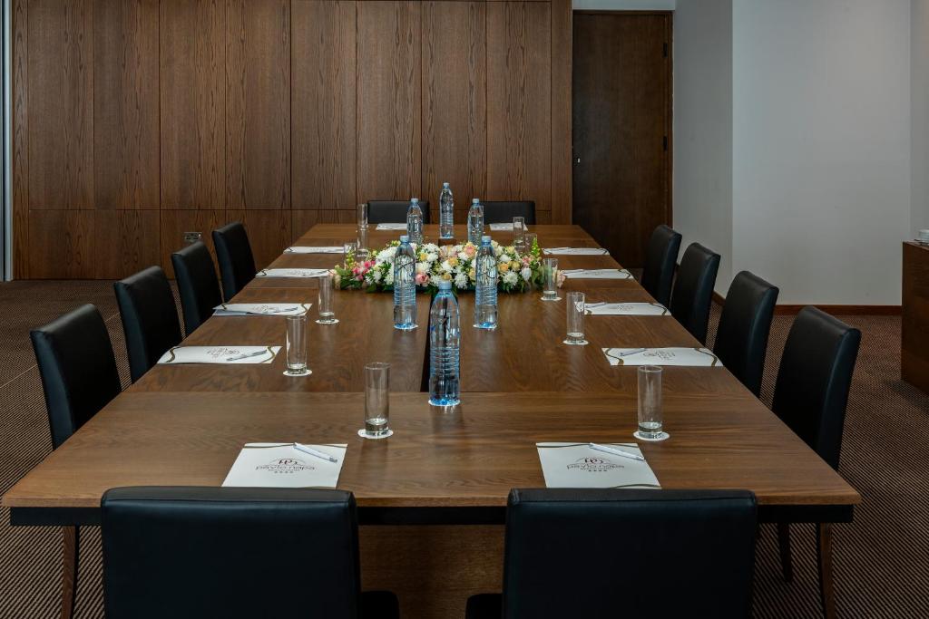 Konferences telpa ar garu koka galdu, melnām ādas krēslām un ziedu kompozīciju vidū. Uz galda novietotas ūdens pudeles un glāzes.