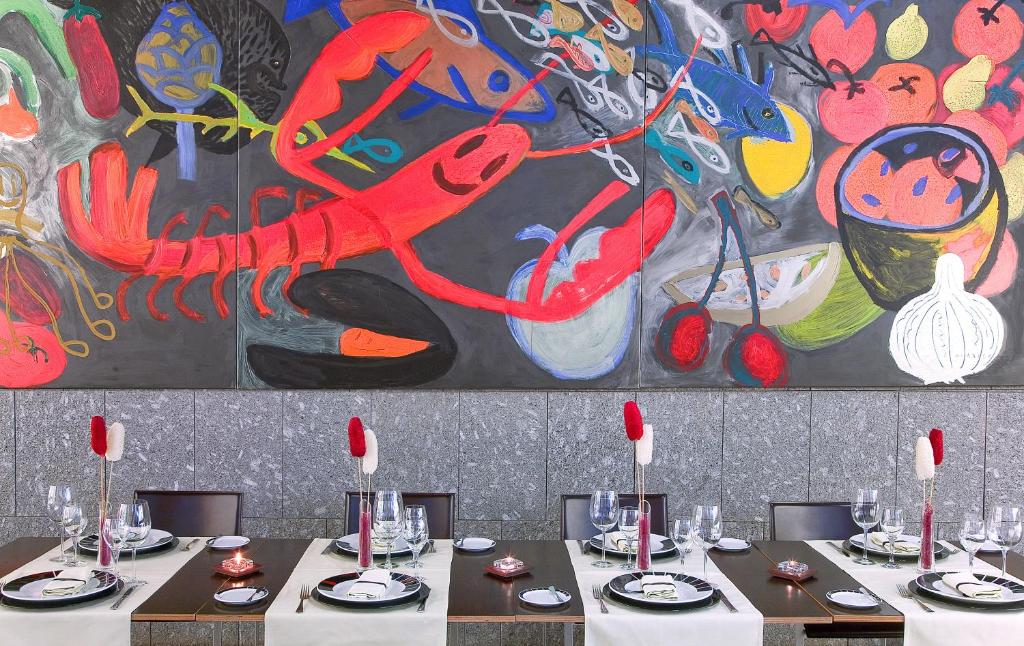 Vibranta mākslas siena ar jūras velšu un dārzeņu motīviem rotā modernu restorānu, kurā galdi ir eleganti uzklāti ar traukiem un glāzēm.