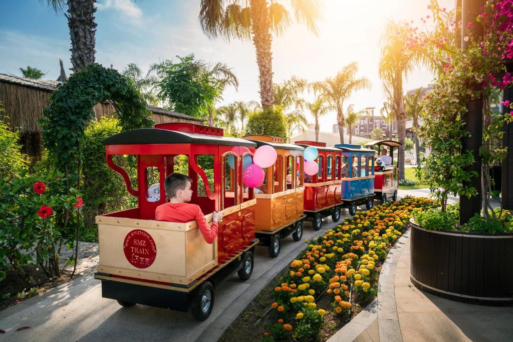 Krāsains bērnu vilcieniņš brauc pa ziedu apstādījumiem, ar palmām un saulainu debesi fona. Bērns sēž vilcieniņa aizmugurē ar baloniem rokās.