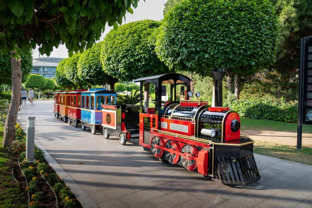 Krāsains mini vilciens brauc pa celiņu, ko ieskauj zaļi koku krūmi. Apkārtējā vide ir mierīga un sakopta, radot patīkamu atmosfēru.