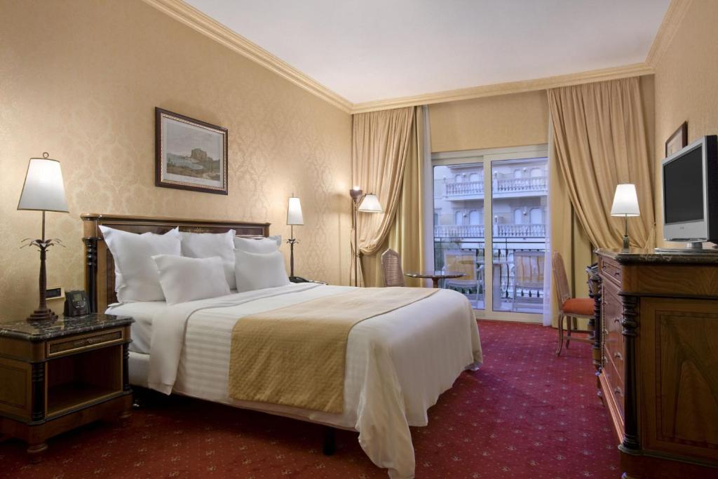 Elegants viesnīcas numurs ar divguļamo gultu, dīvānu un rakstāmgaldu. Labi apgaismots ar lieliem logiem un zeltainām aizkariem, kas rada mājīgu atmosfēru.