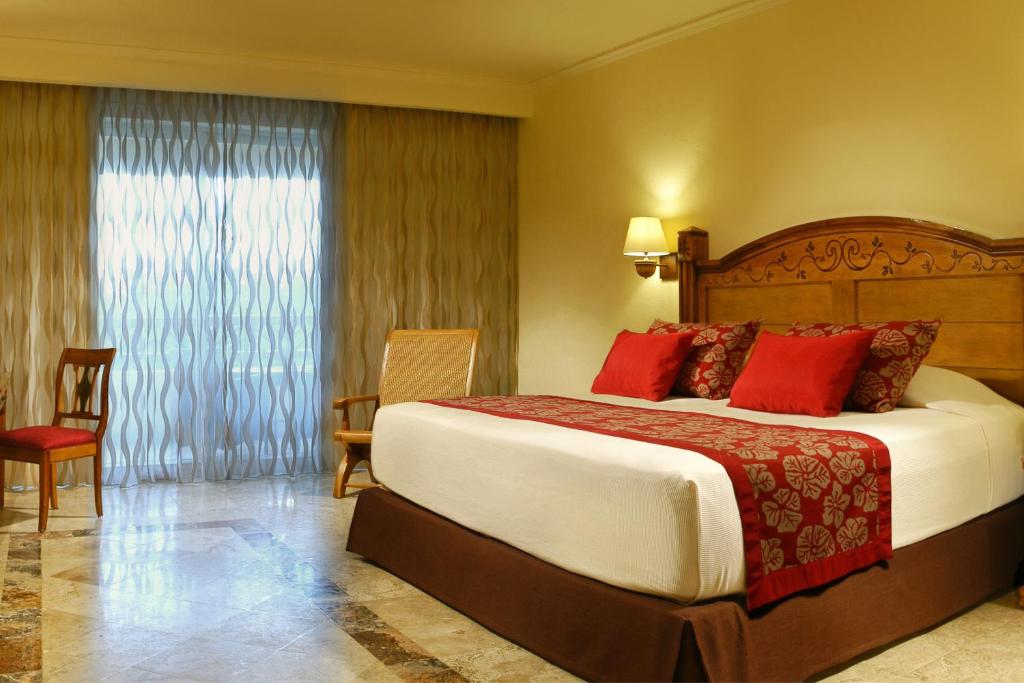 Mūsdienīgs viesnīcas numurs ar plašu gultu, sarkaniem spilveniem un elegantu interjeru, kurā ir arī koka krēsli un caurspīdīgas aizkari.