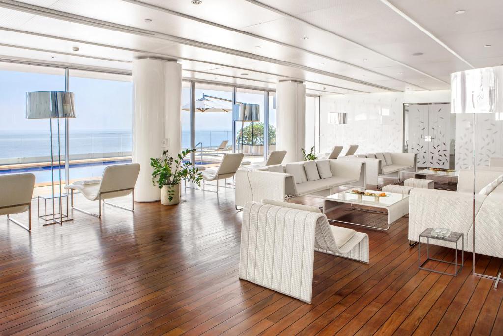 Mūsdienīgs atpūtas telpas interjers ar baltām mēbelēm, koka grīdu un plašu logu, caur kuru redzama jūra. Telpā ir lieli logi, kas nodrošina dabisko apgaismojumu.