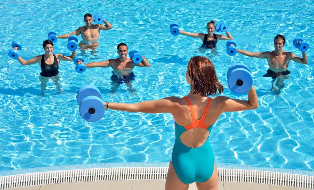 Grupas cilvēku nodarbības ūdens aerobikā, izmantojot zilos ūdens hanteles. Visi dalībnieki ir peldbaseinā un izskatās aktīvi un iesaistīti.