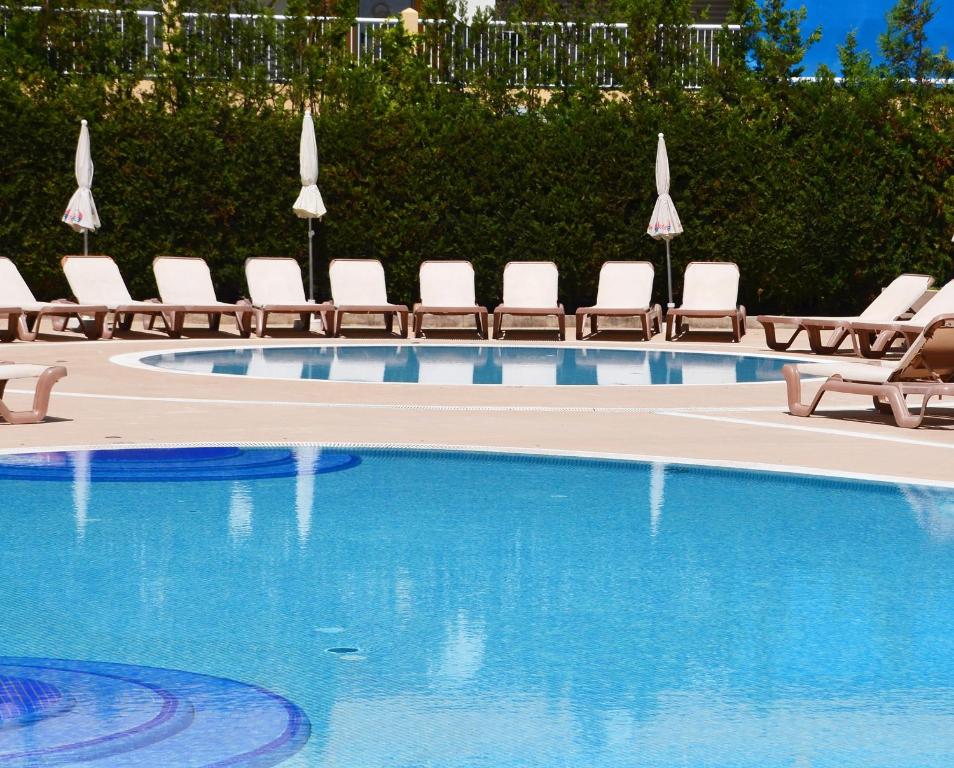 Baseins ar saulessargiem un atpūtas krēsliem, kas izvietoti ap to, radot relaksējošu atmosfēru. Skats uz skaidru ūdeni un apkārtējo zaļo augoņu.