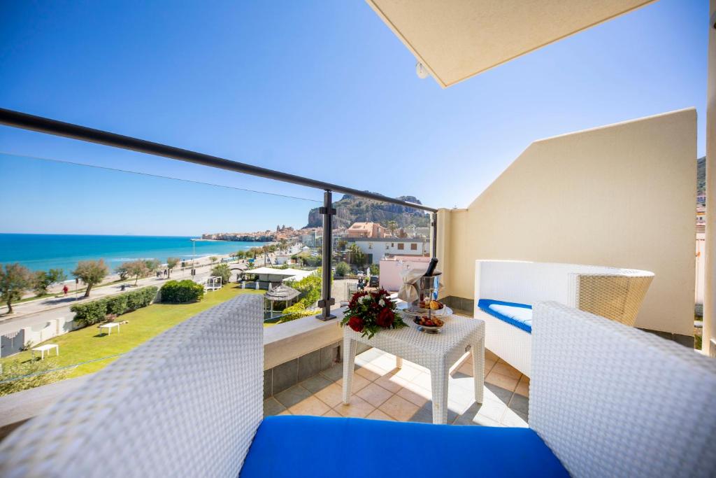 Balkons ar skatu uz jūru, dekorēts ar ērtām baltām mēbelēm un gardumiem, kas nodrošina relaksējošu atmosfēru. Dabiskais apgaismojums un zila debess piešķir ainavai īpašu burvību.