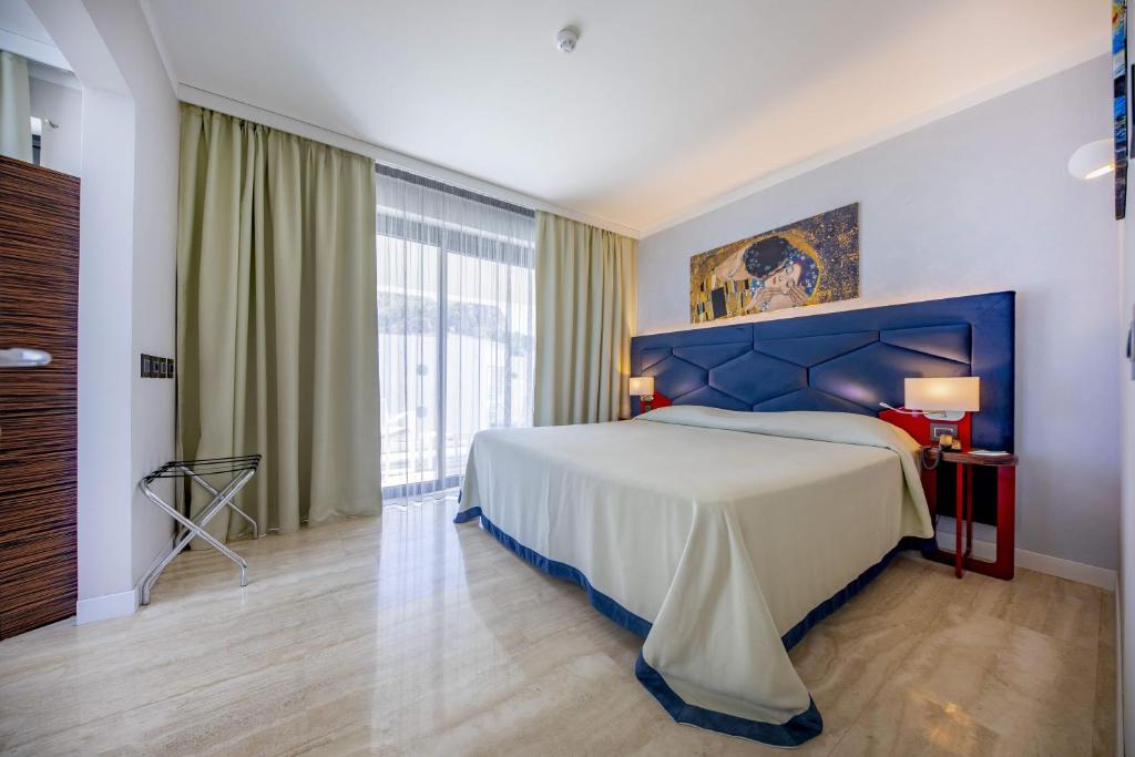 Mūsdienīga guļamistaba ar plašu gultu un stilīgu dekoru, ieskauta ar gaišām aizkariem un ar mākslas darbu uz sienas. Guļamistabā ir dabīga gaisma un elegantas mēbeles.