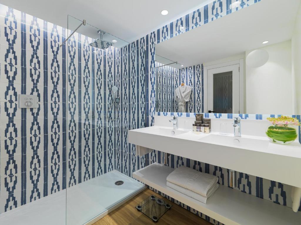 Moderns vannas istabas dizains ar zilo un baltu flīžu rakstu, stikla dušu un elegantu izlietni. Vannas istabā ir arī plaukti un ziedu kompozīcija.