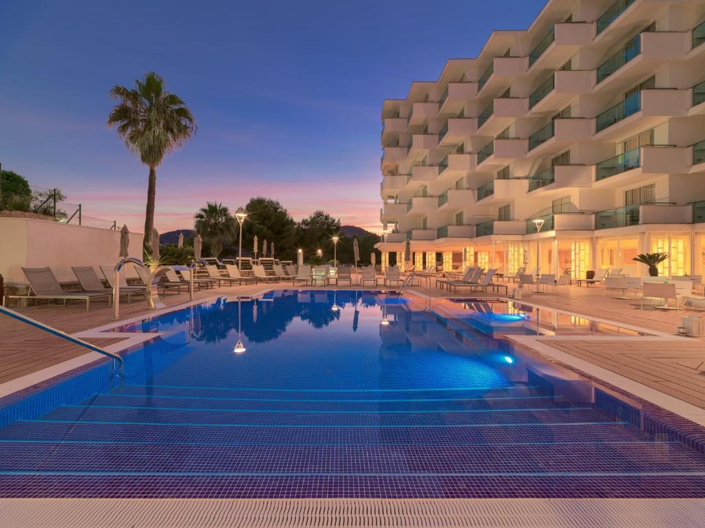 Mūsdienīgs viesnīcu komplekss ar baseinu, ko ieskauj saule un palmas, radot relaksējošu atmosfēru. Vakarā redzams krāšņs saulriets, kas papildina ainavu.
