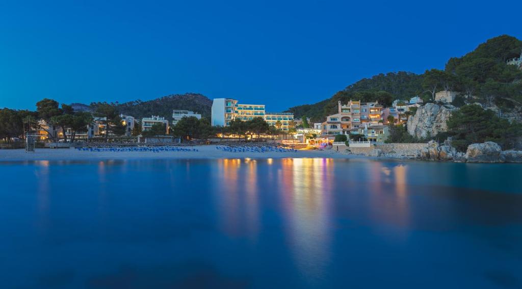 Jūras piekrastes skats ar apgaismotām viesnīcām un pludmali, kurā redzami sauļošanās krēsli un kalnu fons. Mierīga nakts ainava ar atspulgiem ūdenī.