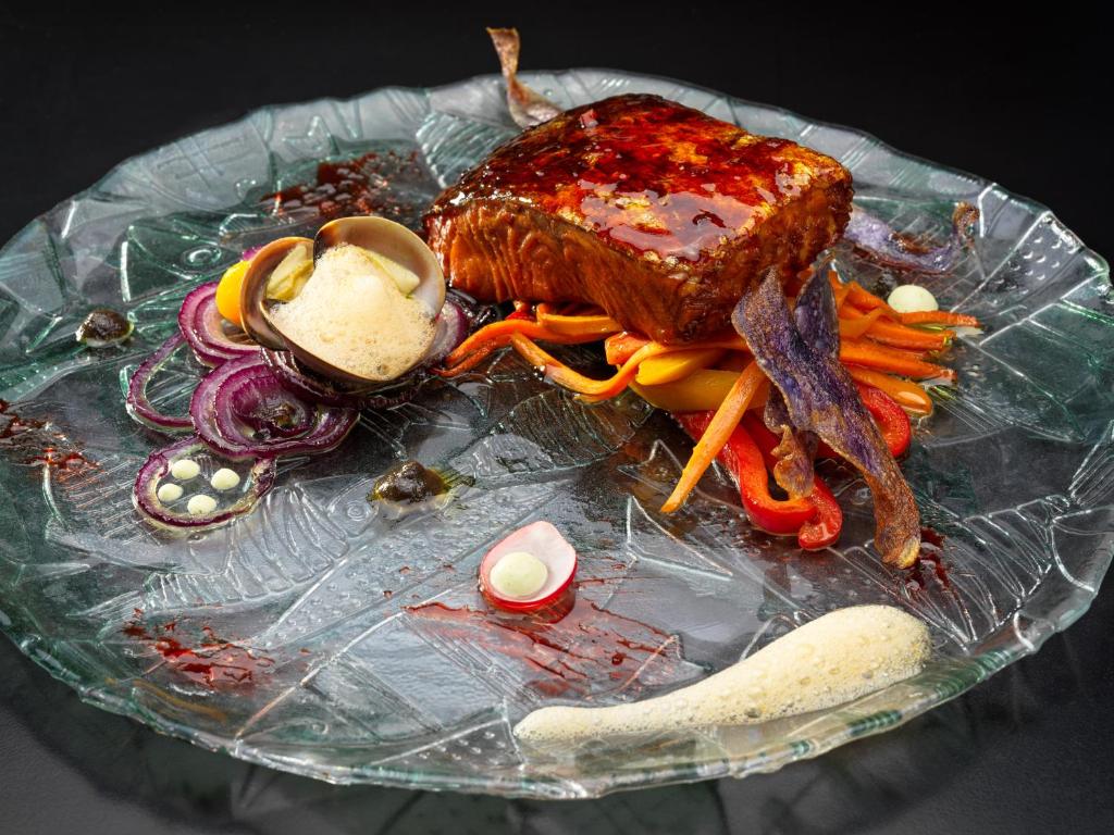 Mūsdienīgs kulinārijas mākslas darbs ar glazētu zivju fileju, kas pasniegta uz krāsainu dārzeņu gultas, papildināta ar garšaugu putu un dekoratīviem dārzeņu elementiem.