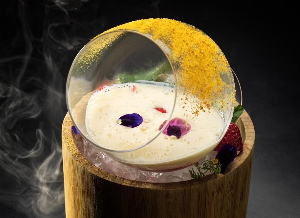 Mūsdienīgs kokteilis pasniegts ar ziedlapiņām un dekoratīvu putu, kas atrodas uz ledus un koka pamatnes.