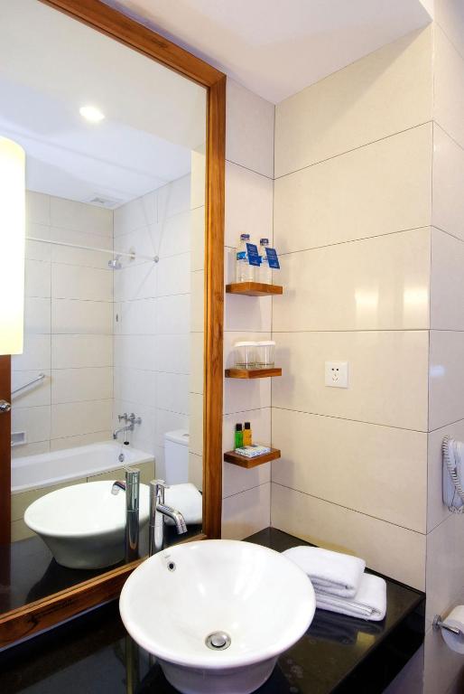 Mūsdienīgs vannas istabas interjers ar baltu izlietni, lielu spoguli un plauktu, kurā atrodas šampūni un citi higiēnas piederumi. Vannas istaba ir gaiša un eleganta, ar melnu akmens virsmu un koka akcentiem.